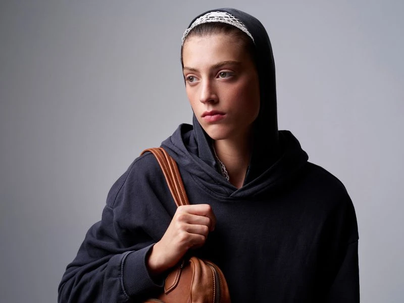Portrait of an adult female wearing a dark hoodie with a white headband, looking away from the camera, and carrying a brown leather bag.