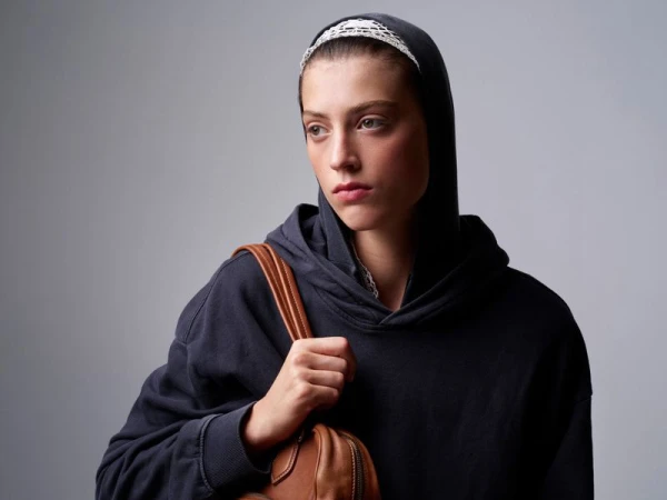 Portrait of an adult female wearing a dark hoodie with a white headband, looking away from the camera, and carrying a brown leather bag.