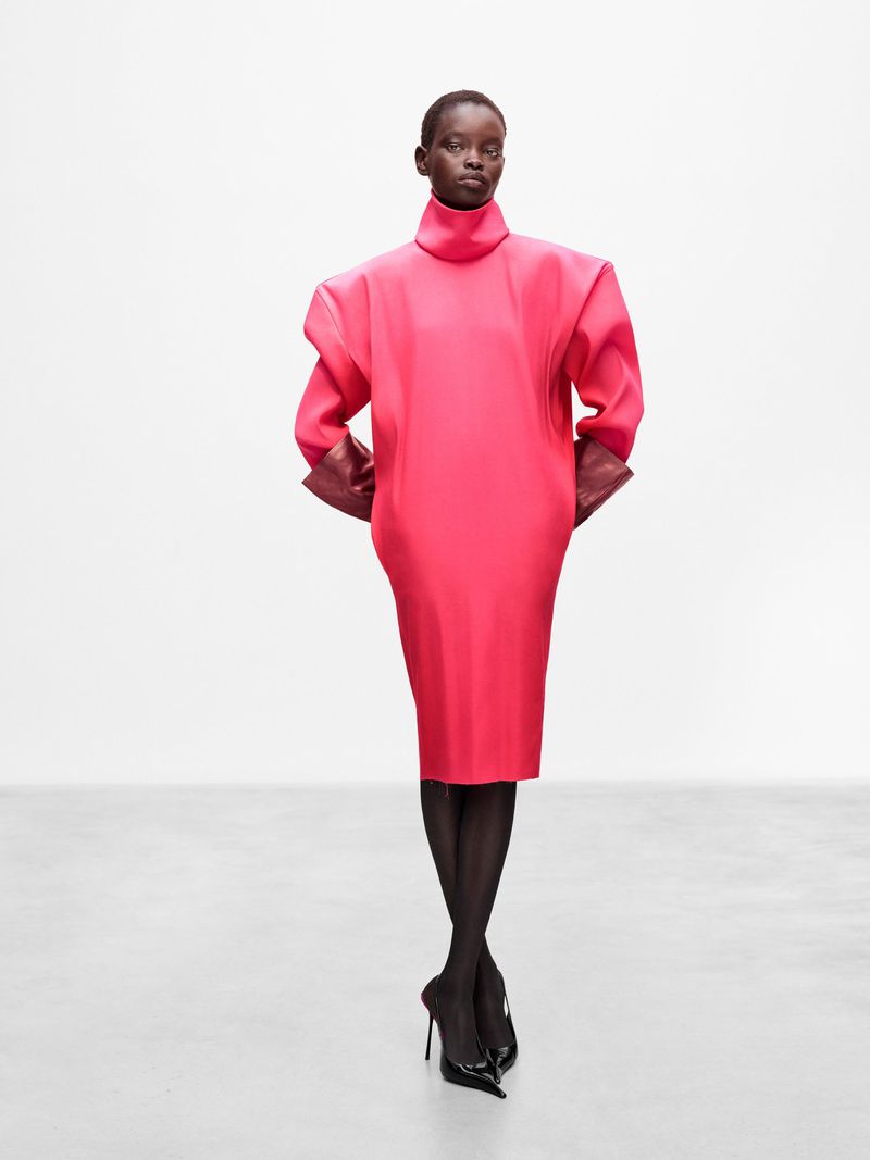 Dark-skinned woman in a vivid pink dress poses against a white background, showcasing an elegant and fashionable attire with black heels.