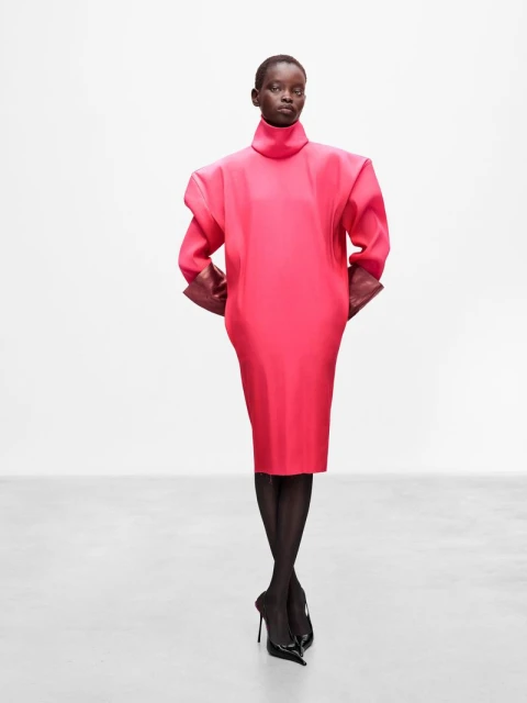 Dark-skinned woman in a vivid pink dress poses against a white background, showcasing an elegant and fashionable attire with black heels.