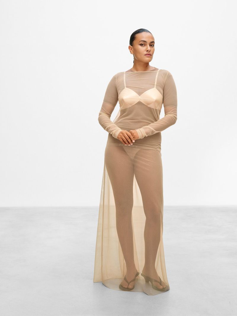 A slender adult female stands in a photo studio, wearing a sheer tan dress and sandals against a bright white backdrop.