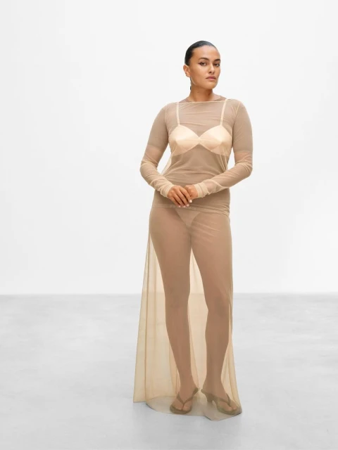 A slender adult female stands in a photo studio, wearing a sheer tan dress and sandals against a bright white backdrop.