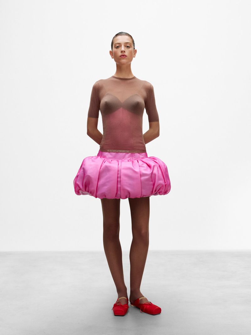 Full shot of a slender woman in a semi-sheer top and puffy pink skirt, hands behind her back, posing against a plain white background.