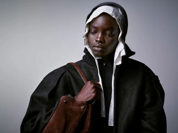 Fashion portrait of a person in a black jacket and white bonnet holding a brown purse against a gray background in studio lighting.