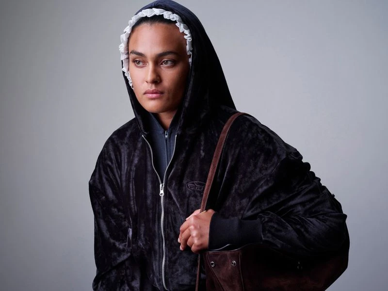 Portrait of a person wearing a black, velvet hooded jacket and a white, ruffled bonnet, carrying a brown suede tote bag, studio setting.