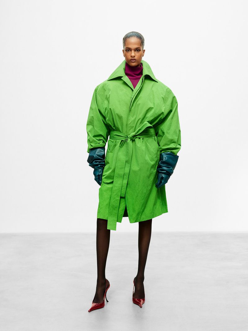Model wearing a bright green belted coat with dark gloves and heels standing against a neutral studio backdrop.