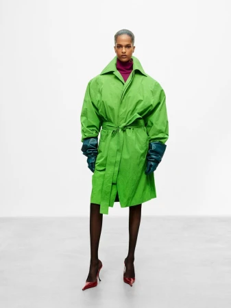 Model wearing a bright green belted coat with dark gloves and heels standing against a neutral studio backdrop.