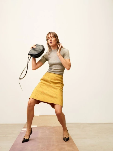 A woman in a yellow skirt and grey top holds a black purse in a studio with a neutral background and a runway-like floor.