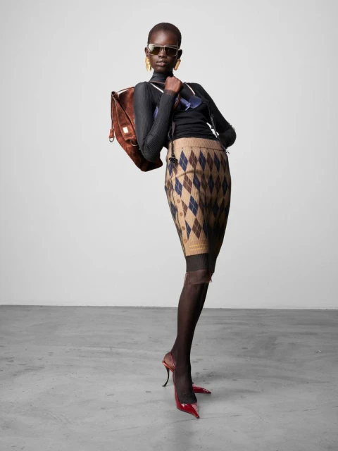 Fashionable woman poses in argyle skirt, heels, and sunglasses with a backpack against a neutral backdrop in a fashion editorial.