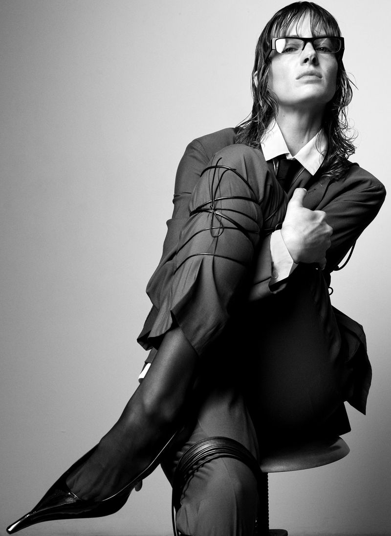 Monochrome portrait of a woman with wet hair wearing glasses and a business suit, seated on a stool in a photo studio against gray background.