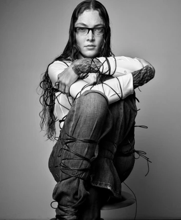 Black and white portrait of a woman with long hair and glasses, sitting on a stool, wrapped in black wires, against a grey background.