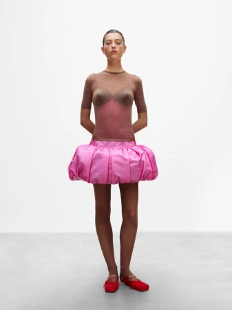 Model in a sheer brown top, voluminous pink bubble skirt, and red ballet flats standing against a plain white studio background.