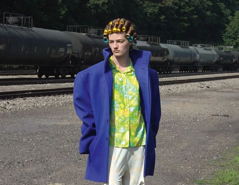 Adult poses outdoors wearing hair curlers, blue coat and tie dye shirt near a train. The shot features a unique and eye-catching fashion style.