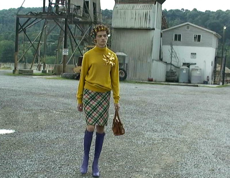 Adult in a yellow sweater and plaid skirt stands in an outdoor industrial gravel lot on an overcast day near a building structure.