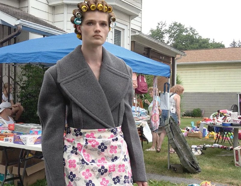 Woman in curlers and a gray coat standing in front of a yard sale, mixing high fashion with everyday life in a suburban setting.