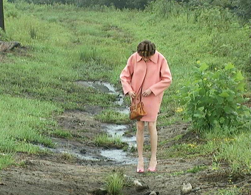 Woman in a pink coat stands on a muddy path in a grassy field, carrying a handbag. Overcast lighting gives the scene a muted, soft look.