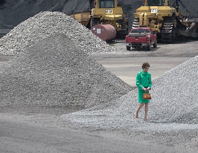 Woman in a green suit walking on a pile of rocks near construction vehicles. She carries a handbag and wears heels.