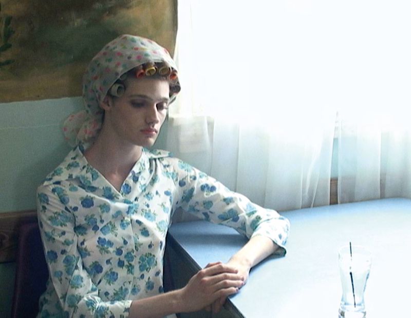 Indoor portrait of a pensive adult woman with hair curlers, floral headscarf and dress, seated at a table next to a glass of water.