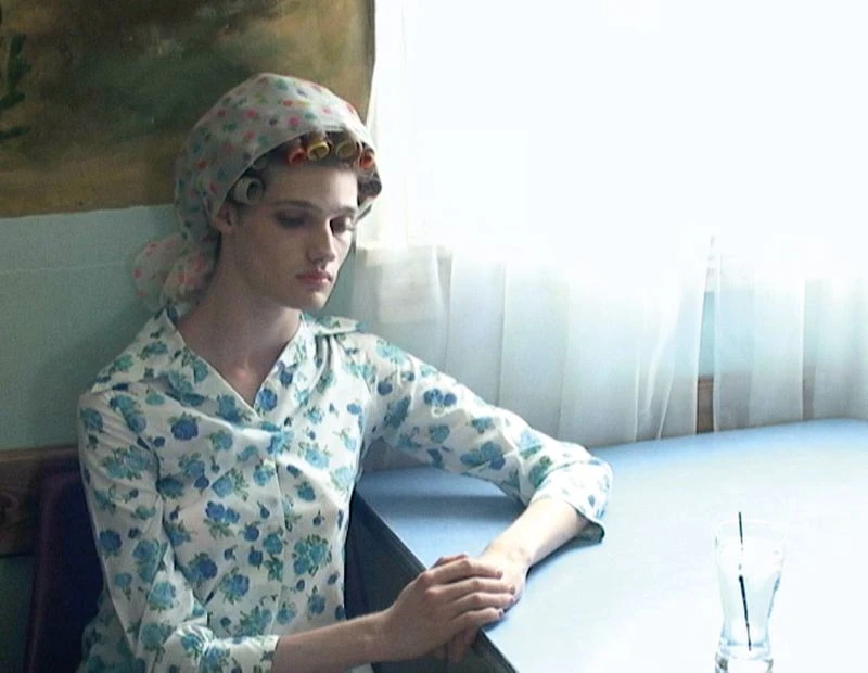 Indoor portrait of a pensive adult woman with hair curlers, floral headscarf and dress, seated at a table next to a glass of water.