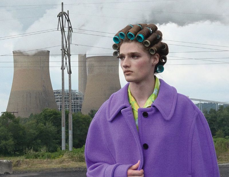 A woman in a purple coat and hair rollers stands in front of a cooling tower and powerlines on an overcast day. Fashion editorial.