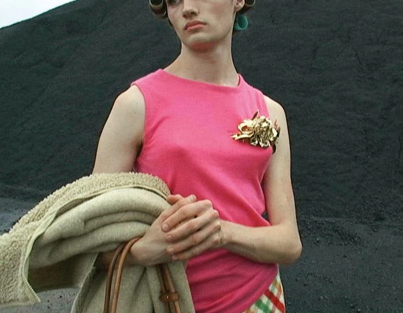 Adult wearing pink tank top, gold brooch, hair rollers, holding jacket against dark background, medium shot. Fashion, portraiture.
