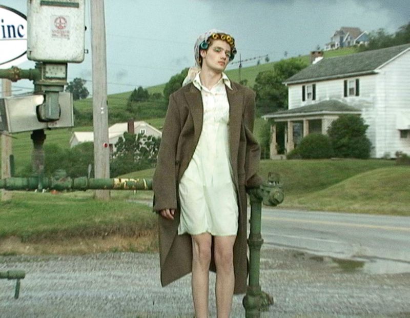 Person in hair rollers, vintage coat and dress standing on street in front of old house and utility infrastructure in a rural setting.