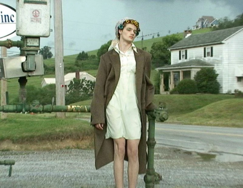 Person in hair rollers, vintage coat and dress standing on street in front of old house and utility infrastructure in a rural setting.