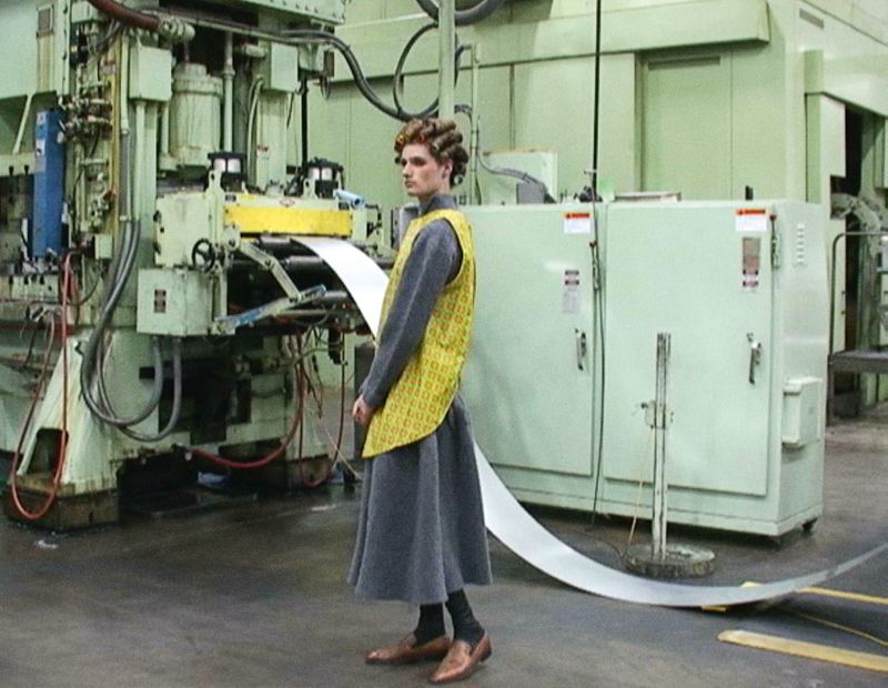 Adult wearing gray dress and yellow vest standing in front of metal machinery in a factory setting, looking off to the side.