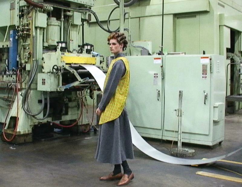 Adult wearing gray dress and yellow vest standing in front of metal machinery in a factory setting, looking off to the side.