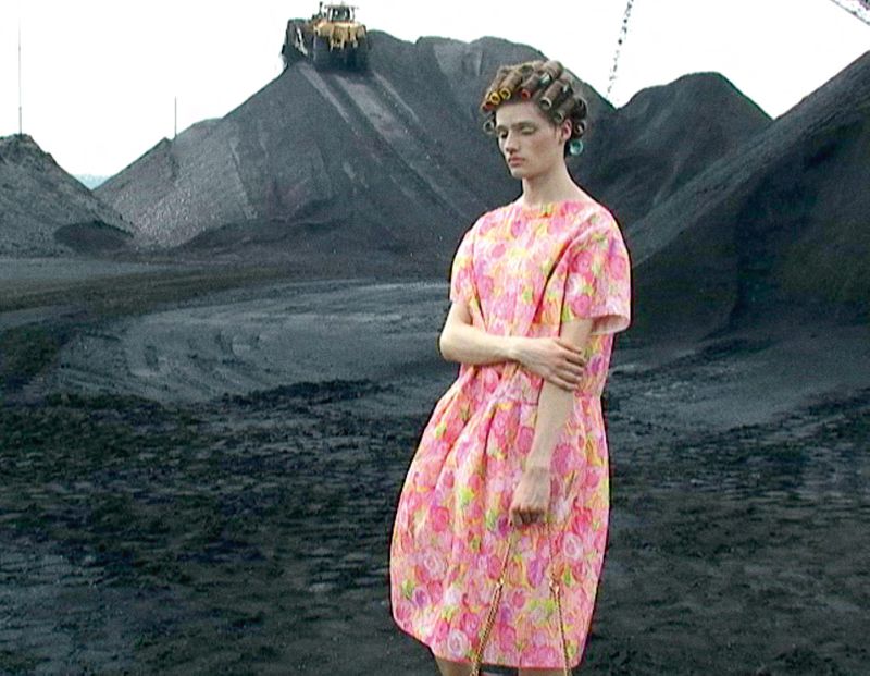 Woman with curlers and pink floral dress stands in a grey, rocky landscape with a construction vehicle driving over a mound.