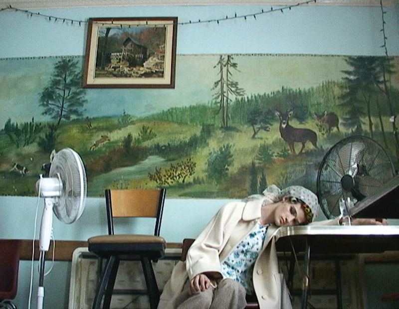 Adult female with blonde hair resting on a table in a room with painted mural, wearing a patterned shirt and white coat, and a head scarf.