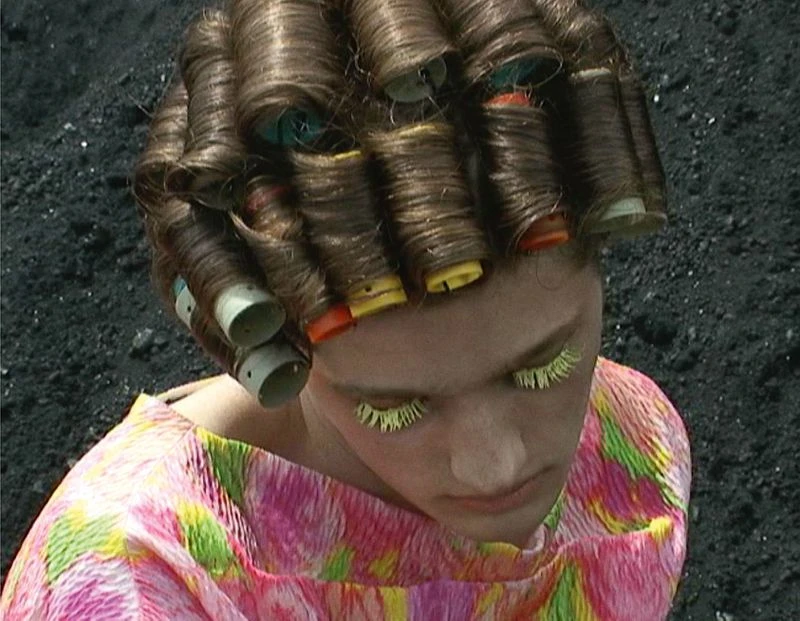 Woman in curlers looks down while standing against a dark background. She wears a multi-colored top and yellow eyelashes.