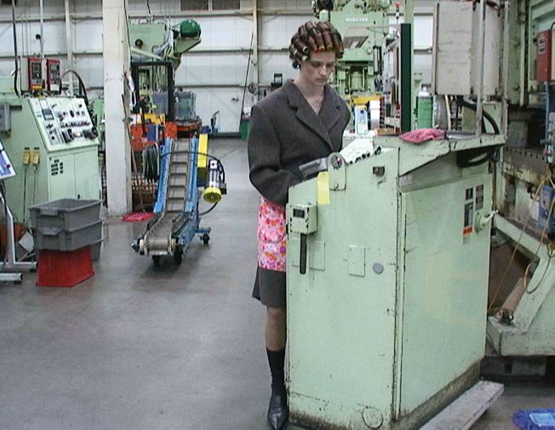 Woman in curlers, apron, and blazer stands in a factory, working at a light green machine with other machinery in the background.