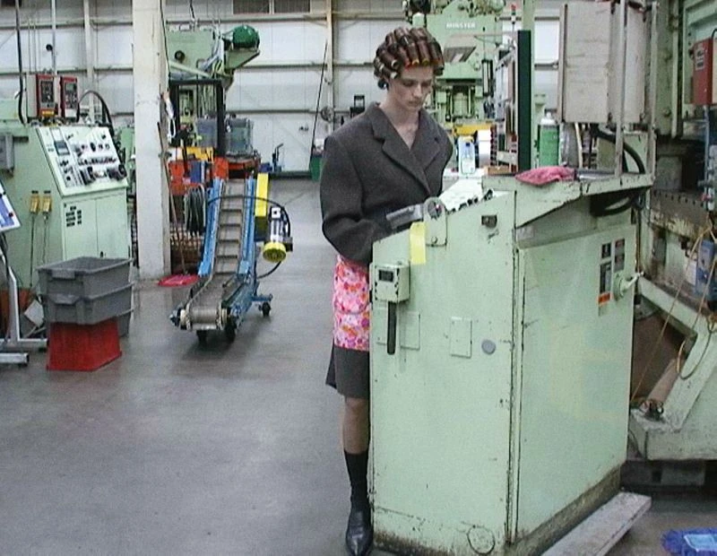 Woman in curlers, apron, and blazer stands in a factory, working at a light green machine with other machinery in the background.