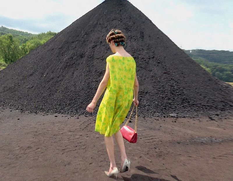 Adult female in a bright yellow dress and hair rollers, walking in an outdoor setting with a pink purse and a large dark hill behind.
