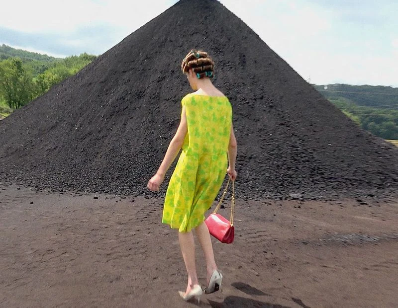 Adult female in a bright yellow dress and hair rollers, walking in an outdoor setting with a pink purse and a large dark hill behind.