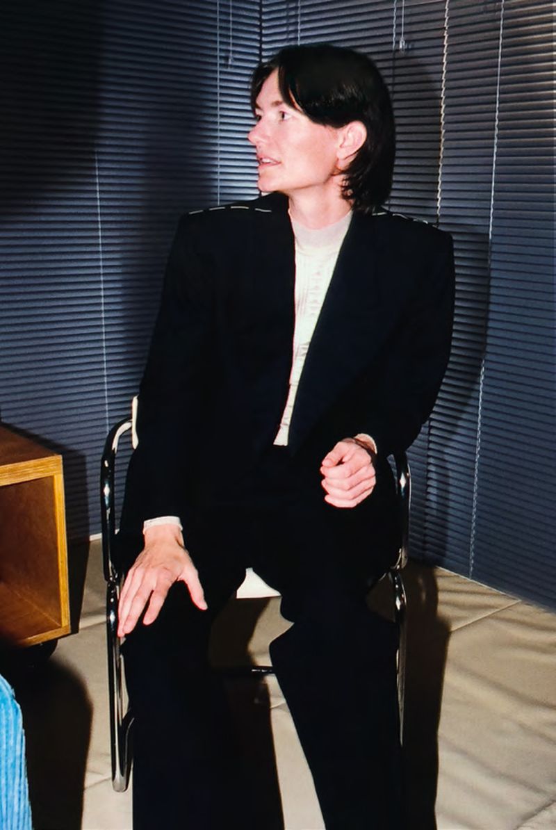 Portrait of an adult wearing a black suit, seated in a minimalist setting, looking off to the side, with window blinds in the background.