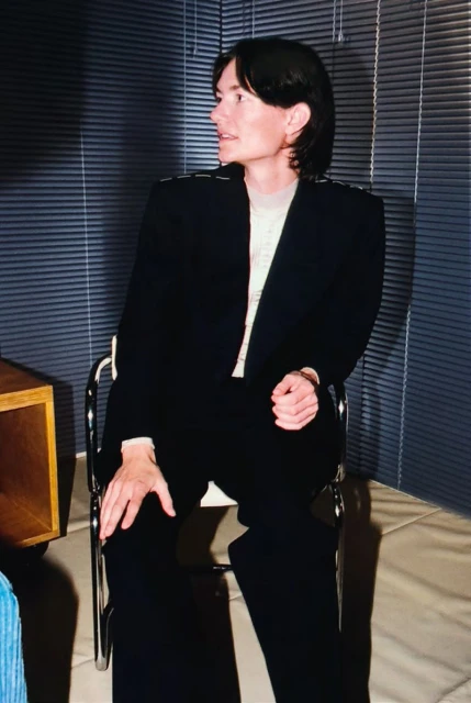 Portrait of an adult wearing a black suit, seated in a minimalist setting, looking off to the side, with window blinds in the background.