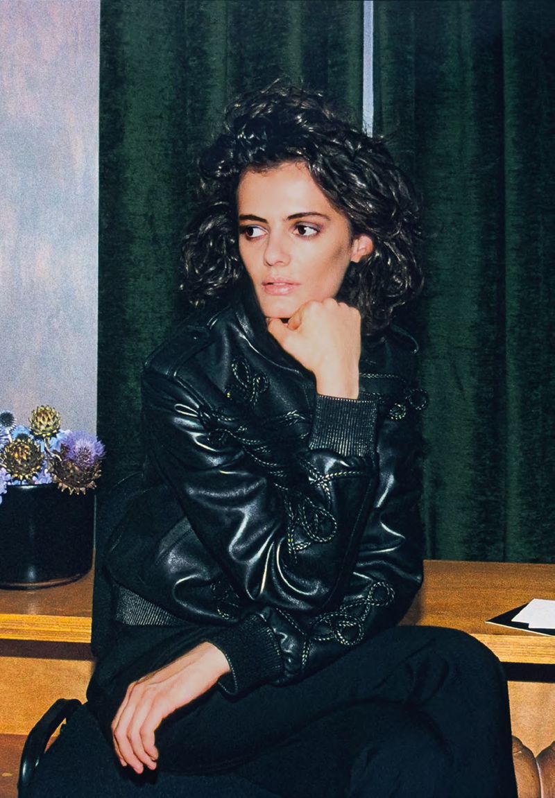 Portrait of a young adult woman with dark curly hair wearing a black leather jacket, seated indoors with a green curtain backdrop.