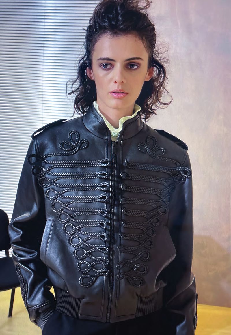 Brunette woman in black leather jacket with decorative frogging detail and yellow collared shirt in front of blinds.