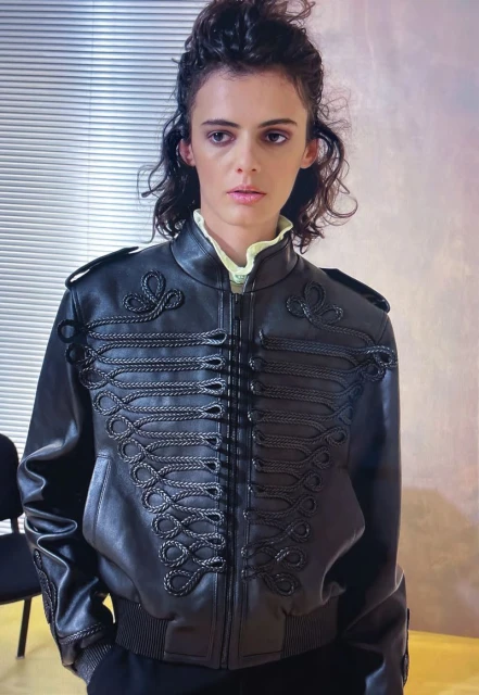 Brunette woman in black leather jacket with decorative frogging detail and yellow collared shirt in front of blinds.