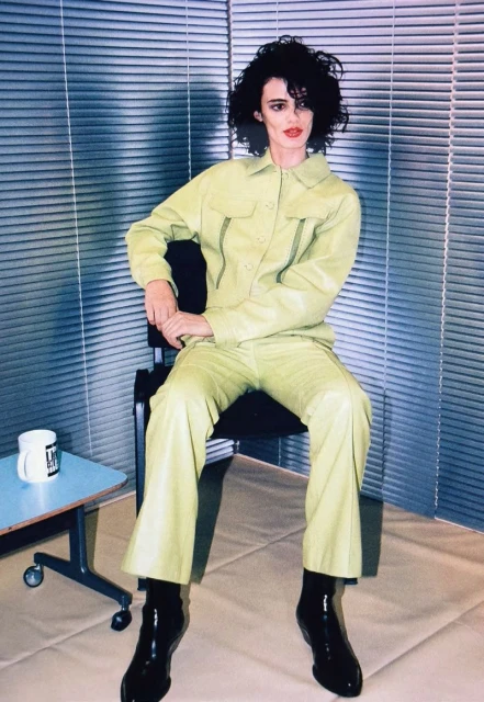 Woman in a green pantsuit sits in front of blinds; coffee mug on a table. She wears black boots and has dark, curly hair and red lipstick.