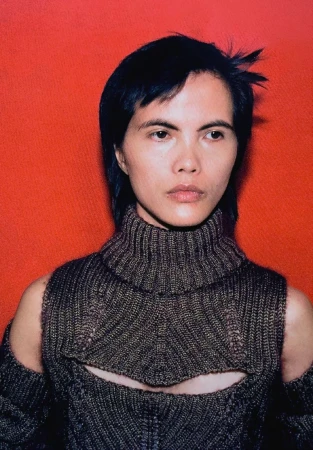 Close-up studio portrait of a young adult with short dark hair wearing a knit sweater against a red background.