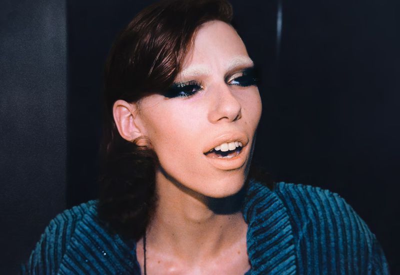 Close-up portrait of adult woman with heavy makeup and a striped turquoise corduroy jacket, facing right against a dark background.