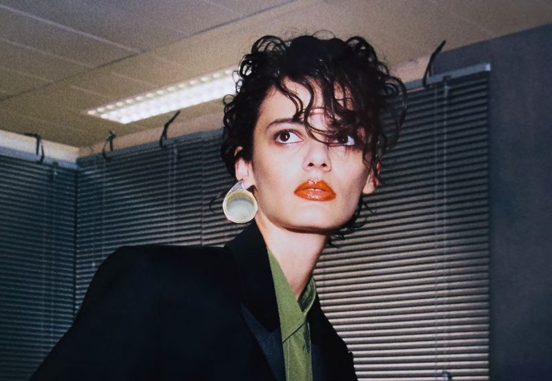 A stylish adult female poses indoors wearing a black blazer, earrings, and bright lipstick, with a neutral expression, in a medium shot.