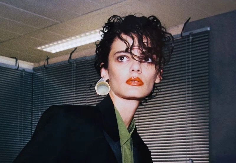 A stylish adult female poses indoors wearing a black blazer, earrings, and bright lipstick, with a neutral expression, in a medium shot.