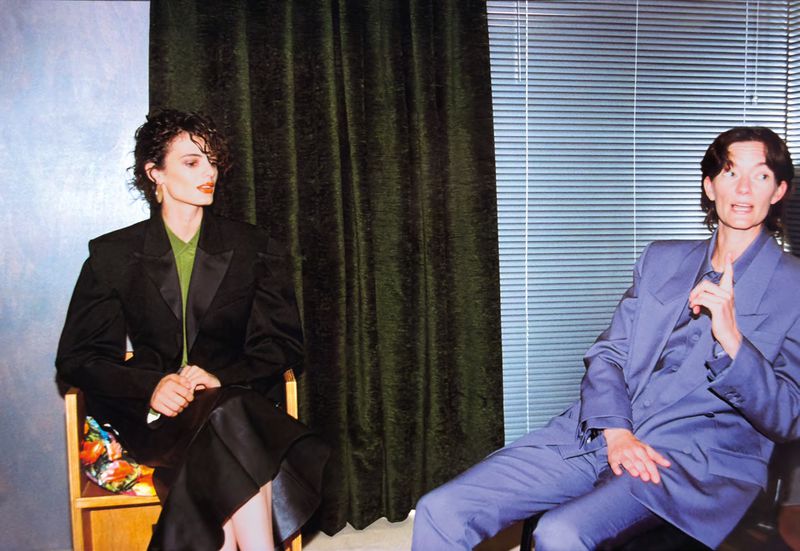 Two adult women in suits seated indoors, one in black, the other in blue, near a window and a green curtain.