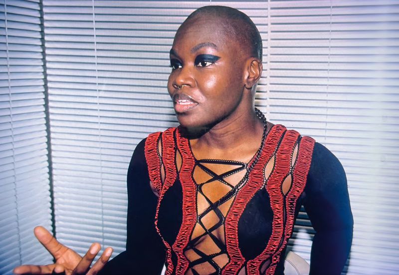 Portrait of Black woman with shaved head and dark eye makeup, wearing a black and orange top, seated against a white blinds background.