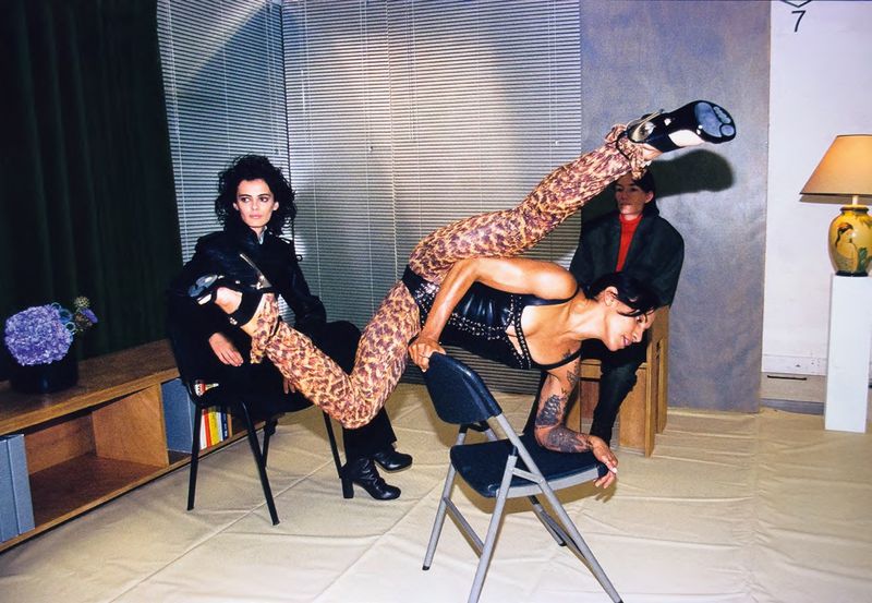 Three women pose in a studio, one balancing on folding chairs in a leopard-print catsuit and black studded top. Fashion editorial.