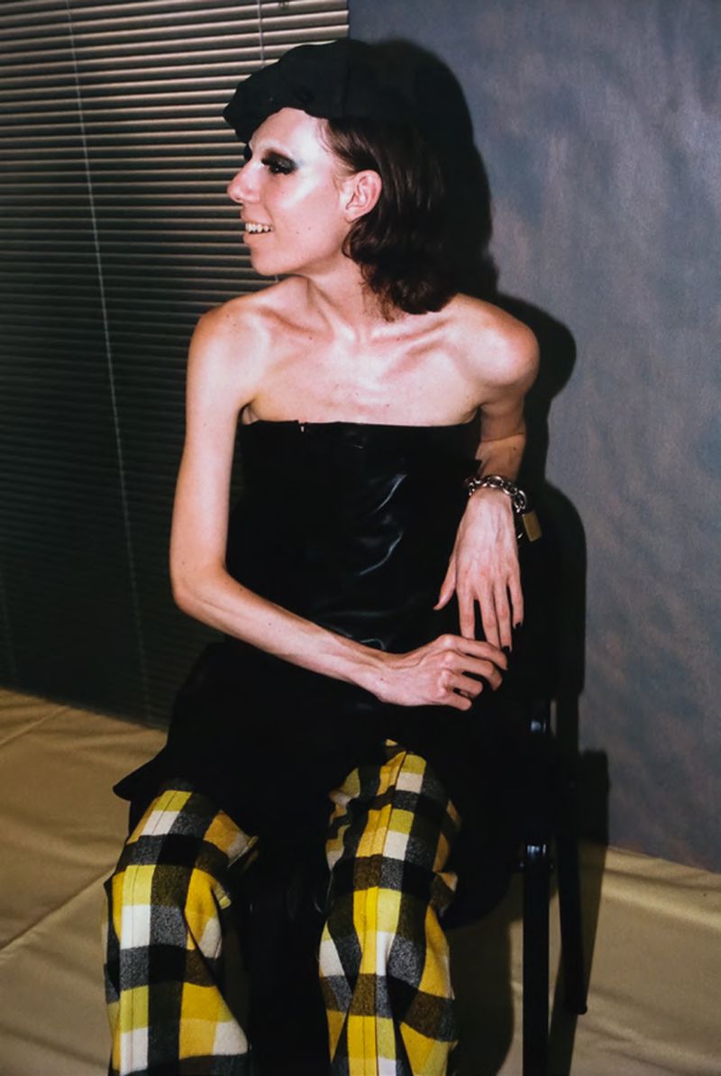 Stylish adult woman in a black strapless top and yellow plaid pants sitting in front of a window with horizontal blinds.